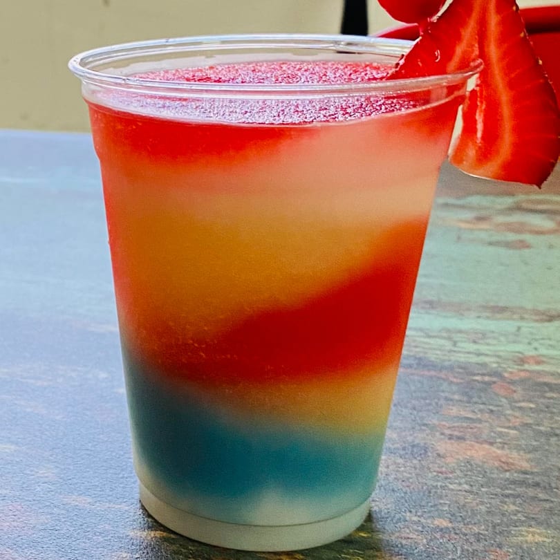 A colorful layered frozen daiquiri in a clear cup, garnished with a strawberry slice.